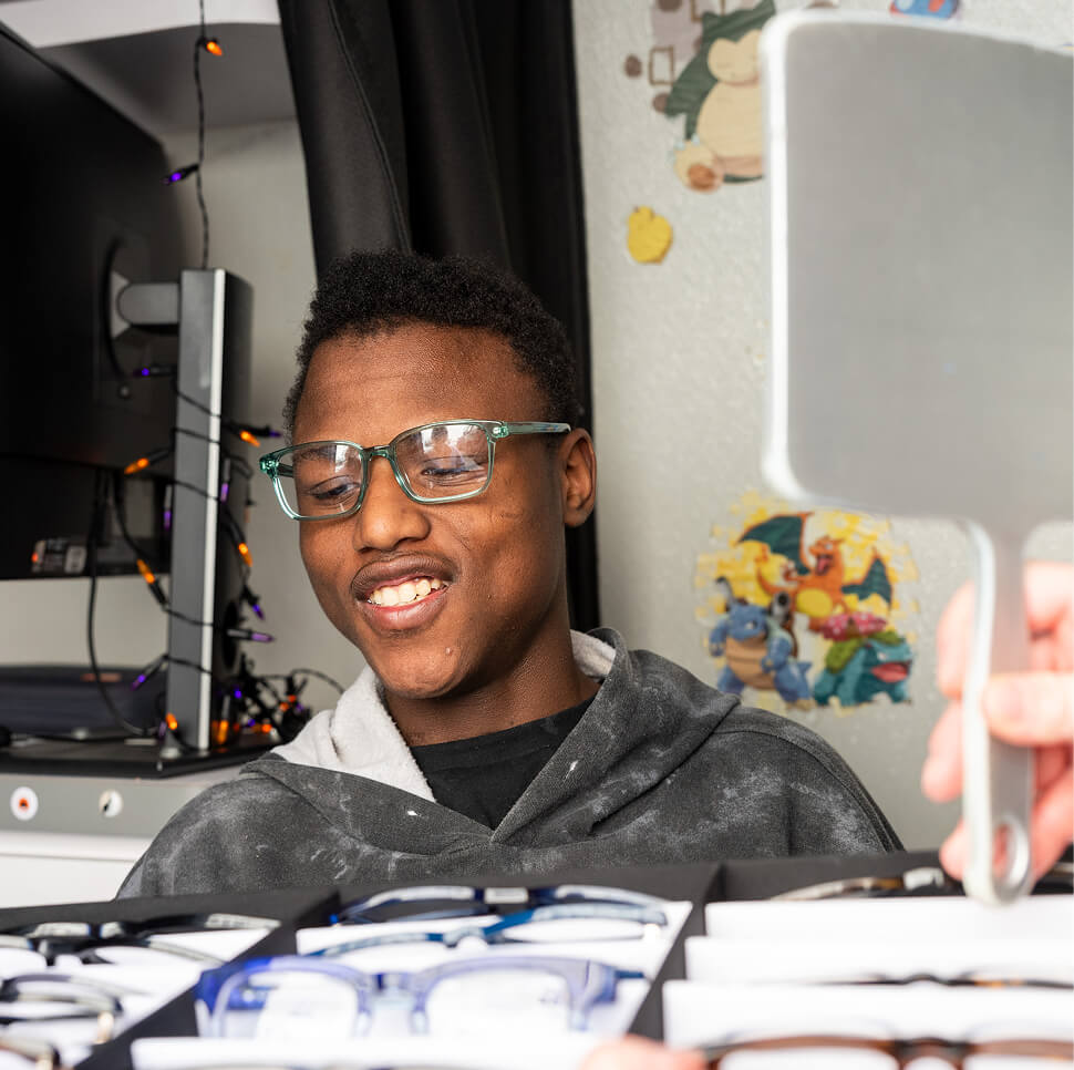 A person wearing glasses looks down at a display of eye glasses while someone holds up a mirror to them.