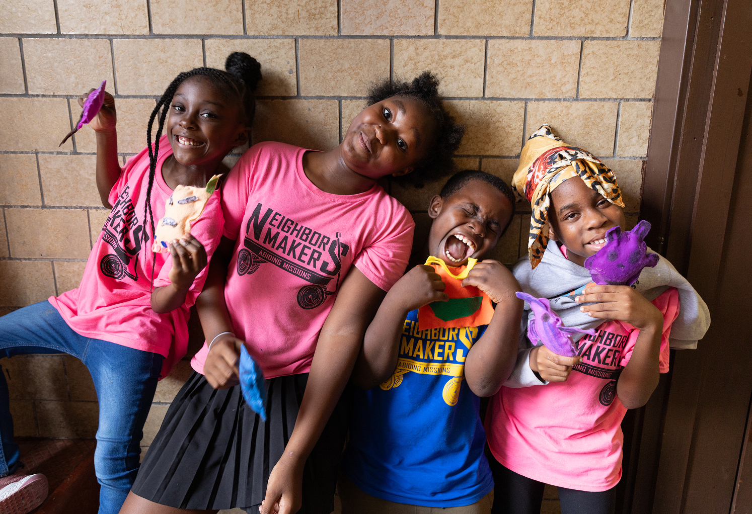 A group of four children wearing Neighbor Makers shirts smile at the viewer.